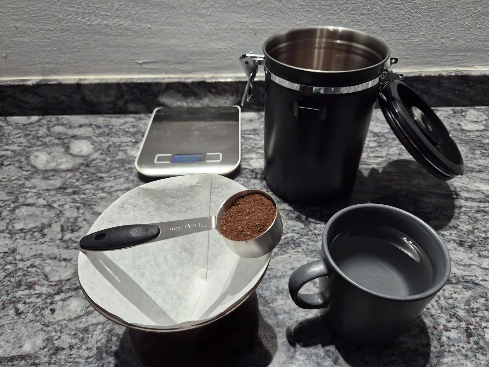 One level spoon of ground coffee next to the paper filters on the V60, the kitchen scale, the coffee tin and a cup filled with water